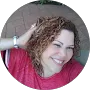 Woman with curly brown hair, smiling, hand in hair, wearing red shirt, outdoors.